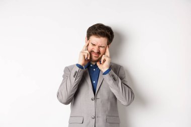 Takım elbiseli bir iş adamının başı ağrıyor, yüzü buruşuyor, başı ağrıyor, migren oluyor, beyaz arka planda hasta gibi duruyor.