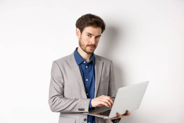 Takım elbiseli yakışıklı iş adamı dizüstü bilgisayarda çalışıyor, kameraya özgüvenli bakıyor, beyaz arka plana karşı duruyor.