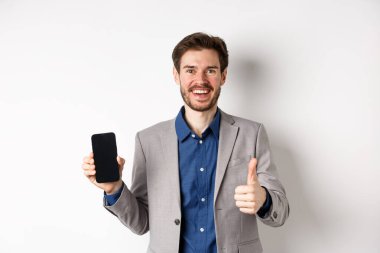 E-ticaret ve online alışveriş konsepti. Takım elbiseli tatmin olmuş iş adamı boş akıllı telefon ekranı ve baş parmak yukarı gösteriyor. İyi bir anlaşma, beyaz arka plan.