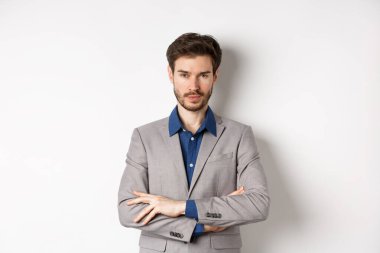 Profesyonel gibi görünen kendine güvenen genç adam, göğsünde çapraz kollar, beyaz arka planda duruyor.