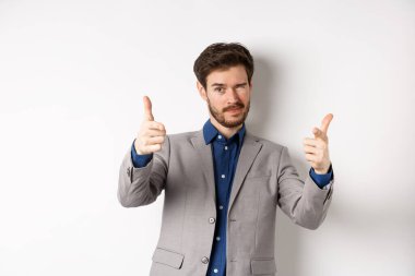 Sen halledersin. Beyaz takım elbiseli yakışıklı beyaz iş adamı kamerayı işaret ediyor ve göz kırpıyor, insanları motive ediyor, şirkette çalışmaya davet ediyor, beyaz arka planda duruyor.