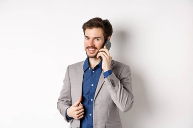 Başarılı iş adamı takım elbisesini onarıyor ve cep telefonuyla konuşuyor. Kameraya gülümsüyor, beyaz arka planda duruyor.