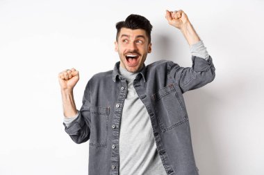 Heyecanlı genç adam tezahürat yapıyor ve yana bakıyor, ödül piyangosunu kazanıyor, iyi bir anlaşma yapıyor ve kutluyor, amacına ulaşıyor veya başarıyor, zafer dansı yapıyor ve evet diye bağırıyor, beyaz arka plan