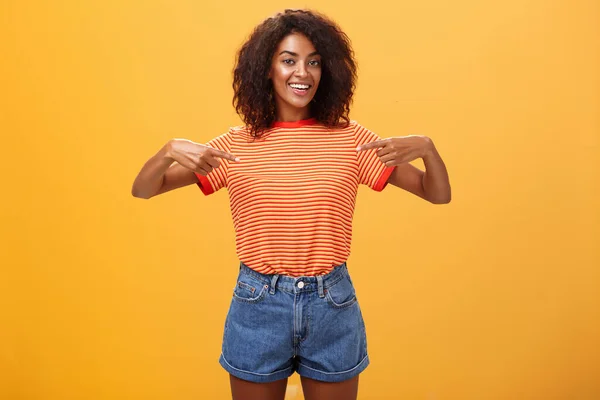 Hey pick me I your girl looking for. Portrait of charming friendly-looking ambitious dark-skinned female with afro hairstyle pointing at chest proudly and joyful posing against orange background