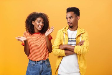 Guy thinks his friend weirdo making dumb thinks looking at cute girl with suspicious look crossing arms on chest raising eyebrow questioned while girlfriend saying sorry shrugging over orange wall