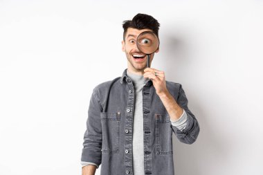 Mutlu komik adam büyüteçten büyük gözlerle bakıyor, ilginç bir anlaşmaya bakıyor, bir şeyler arıyor, beyaz arka planda duruyor.