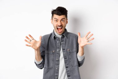 Kızgın adam çığlık atıyor ve kameraya bakıyor, öfkeli ve nefret dolu bir ifadeyle bağırıyor, beyaz arka planda duruyor.