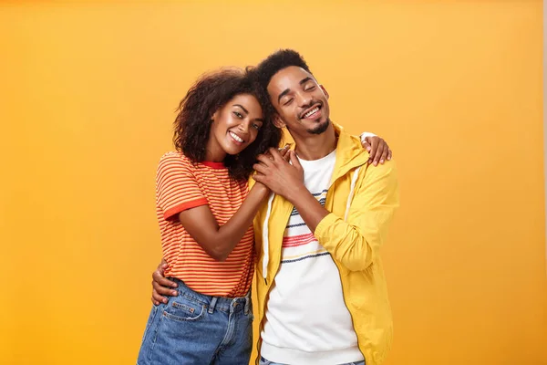 They love each other. Portrait of two charming african american man and woman in relationship hugging with heartwarming smile touching hands smiling gently posing against orange background