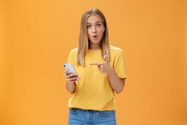 Akıllı telefonlara bağlanan kablosuz kulaklıklardan etkilenen adam ekranı işaret ederek sorgulanıyor ve şaşırtılan kıvrılmış dudaklar turuncu duvarın üzerinde kulaklık takan kameraya bakıyor.