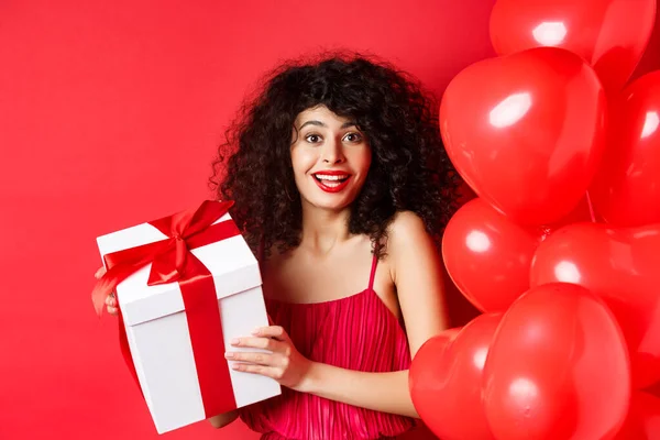 Holidays and celebration. Beautiful woman with curly hair, standing near heart balloons, holding gift box and smiling happy, white background