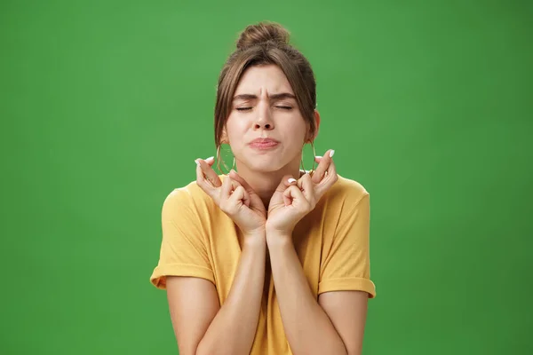 Preocupado intenso, preocupado atraente jovem modelo feminino europeu em amarelo t-shirt levantando ombros cruzando os dedos para boa sorte bolsa lábios e fechar os olhos enquanto espera por milagre espero — Fotografia de Stock