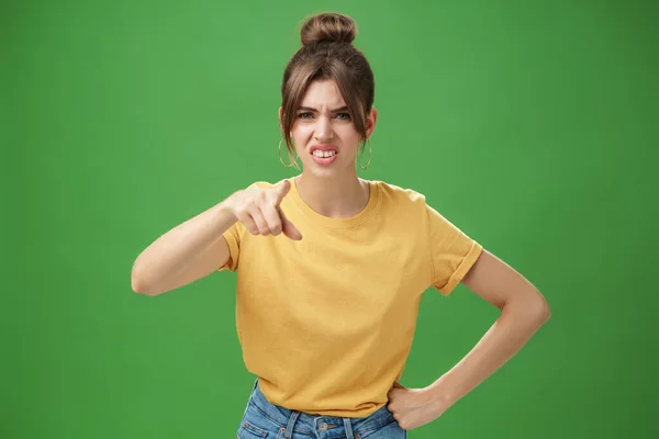 Portrét arogantní neslušné mladé ženy ve žlutém tričku ukazující na kameru, mžourající a šklebící se z pohrdání a opovržení, stojící neohromeně na zeleném pozadí — Stock fotografie