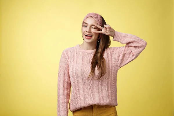 Alegre feliz glamour joven mujer europea en suéter de punto rosa, usando diadema, guiñando un ojo coqueto y lindo, mostrando la victoria o signo de paz sobre el ojo, sintiéndose emocionado y alegre sobre la pared amarilla — Foto de Stock