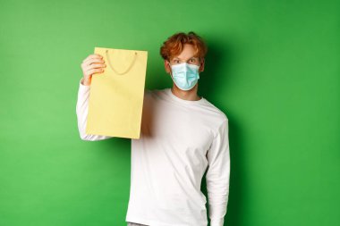 Covid-19 ve pandemik konsept. Yüzü maskeli, alışveriş torbasını gösteren ve yeşil arka planda duran kameraya bakan şaşırmış bir adam.