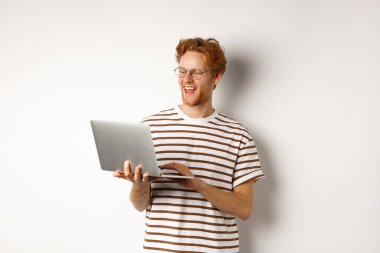 Laptop 'ta serbest çalışan yakışıklı kızıl saçlı adam, gülümsüyor ve bilgisayar ekranına bakıyor, beyaz arka plan.