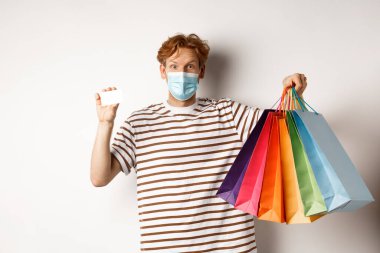 Sosyal uzaklık, koronavirüs konsepti. Kızıl saçlı genç adam, maske takıyor, alışveriş torbaları ve plastik kredi kartı gösteriyor, beyaz arka plan.