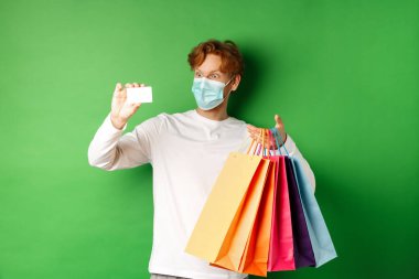 Tıbbi maskeli yakışıklı, plastik kredi kartı ve alışveriş torbaları gösteren indirimli, yeşil arka planlı bir genç.