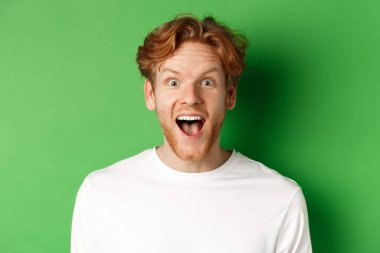 Duygular ve moda konsepti. Şaşırmış kızıl saçlı adama yakın çekim yapmak için kameraya neşeli bir şekilde bakan yeşil arka planda duran harika bir haber var.