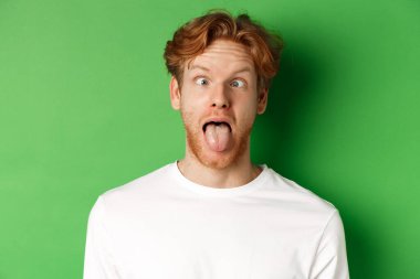 Duygular ve moda konsepti. Komik, kızıl saçlı, aptal suratlar gösteren, dil uzatan ve gözlerini kısan, yeşil arka planda duran adamın yakın çekimi.