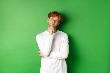 Gözlüklü düşünceli kızıl saçlı adam, seçim yapıyor, yukarı bakıyor ve düşünüyor, yeşil arka planda duruyor.