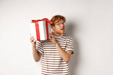 Sevgililer günü ve tatil konsepti. Şu anki kutusunun içinde ne olduğunu tahmin etmeye çalışan ilginç bir genç adam. Kızıl saçlı adam hediyesini kulağının yanında sallıyor, beyaz arkaplan.