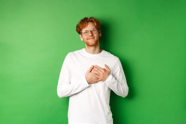 Handsome young man in glasses and white long-sleeve smiling, holding hands on heart with grateful face, thanking you, standing over green background — Stock Photo, Image