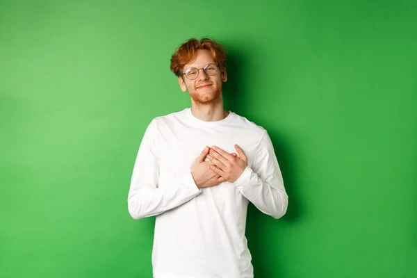 Joven guapo con gafas y manga larga blanca sonriendo, agarrado del corazón con cara agradecida, agradeciéndote, de pie sobre fondo verde — Foto de Stock