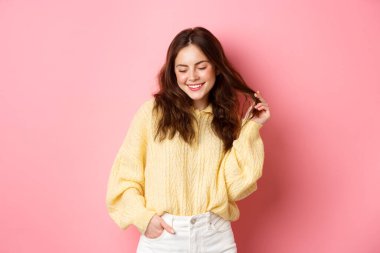 Şirin ve romantik esmer kadın saçlarıyla oynuyor, utangaç bir gülümsemeyle aşağıya bakıyor, pembe arka plana karşı flört ediyor.