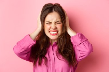 Close up of pissed-off and angry woman grimacing bothered, shut her ears with hands from loud bothering noise, standing over pink background