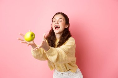 Mutlu tatlı kız bir elma yakalar ve güler, sağlıklı öğle yemeği zamanı meyvelerle dolu, pembe arka planda durur