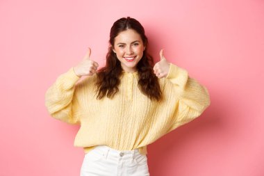 Aferin, çok iyi. Gülümseyen genç bir kadın senin seçimin gibi onaylıyor. İyi ki pembe arka plana karşı tatmin olmuş bir şekilde duruyorsun.