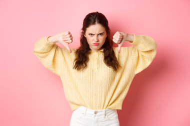 Kızgın ve hoşnutsuz genç bir kız, kötü bir şeye burun kıvırıyor, hoşnutsuzluğunu ifade ediyor, olumsuz tepkiler veriyor, pembe arka plana karşı üzgün duruyor.