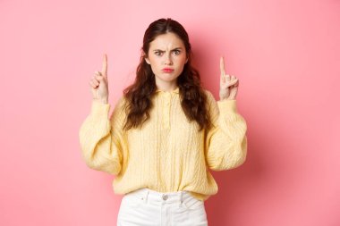 Hüzünlü kasvetli kız şikayet ediyor, kaşlarını çatıyor ve kötü işaret veya logoyu işaret ediyor, bir şeyden memnun değil, pembe arka plana karşı duruyor.