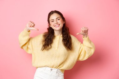 Mutlu gülümseyen, kendini gösteren, kendini tanıtan, kendine güvenen genç bir kadın, pembe arka plana karşı durarak beni seç.