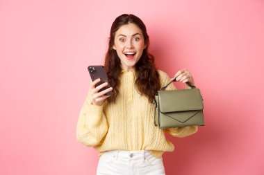 Heyecanlı esmer kız, elinde akıllı telefonuyla online mağazadan çanta sipariş etti. Yeni çantasını gösteriyor ve şaşkın bir şekilde gülümsüyor. Pembe arka planda duruyordu.
