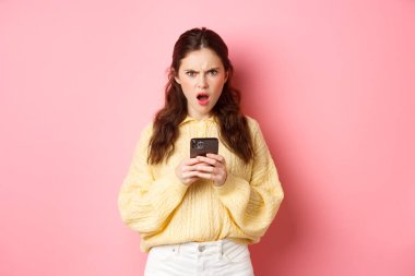 Kafası karışmış ve kızgın kız arkadaş telefonda garip mesajlar okuyor, kameraya bakıyor hayal kırıklığına uğramış ve sorgulanmış, pembe arka plana karşı duruyor.