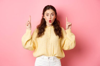 Şaşırmış esmer kız hayret içinde hayret ediyor, hayret ediyor ve kameraya bakıyor. Etkilenmiş, telif alanını işaret ediyor, pembe arka planda duruyor.