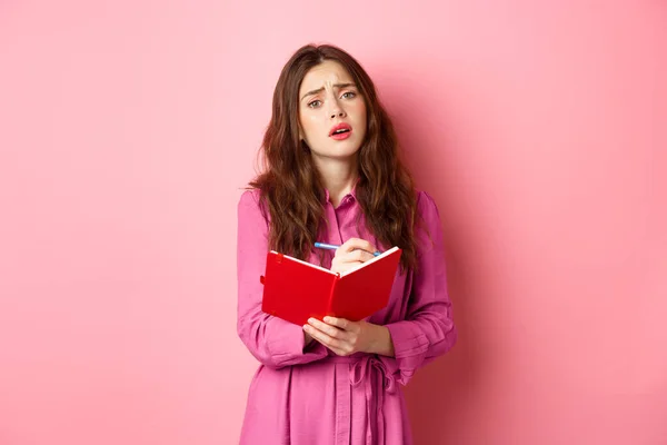 Coy romantic girl sighing, writing in her diary about sad moment, making notes in planner, standing against pink background