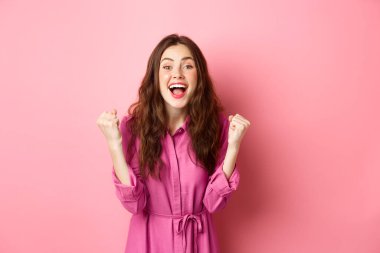 Gülümseyen güzel bir kadın mutlu ve heyecanlı bir yüzle çığlık atıyor, evet diyor, yumruk sallıyor, kazanıyor ve şampiyon gibi hissediyor, zafer kazanıyor, pembe arka planda duruyor.