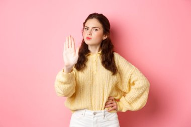 Kızgın ve otoriter genç kadın kaşlarını çatıyor, ciddi görünüyor, engelleyici hareketler sergiliyor, hayır demek için elini uzatıyor, kötü bir şeyi reddediyor, eylemi yasaklıyor, pembe arka plana karşı duruyor.