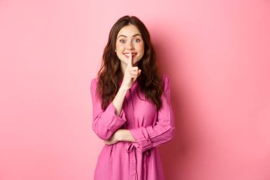 Güzel genç bir kadın gülümsüyor ve şşşş yapıyor, parmaklarını dudaklarına bastırıyor sessiz kalmak ya da gizli kalmak istiyor, pembe arka planda duruyor.