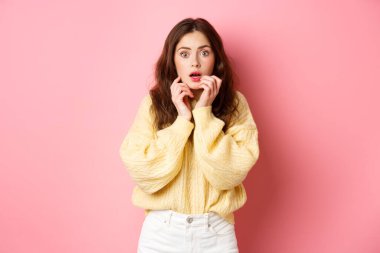 Korkmuş ve şok olmuş esmer bir kızın portresi, nefes nefese ve dudaklarının yanında el ele tutuşmuş, korkmuş bir şekilde kameraya bakıyor, pembe arka planda duruyor.