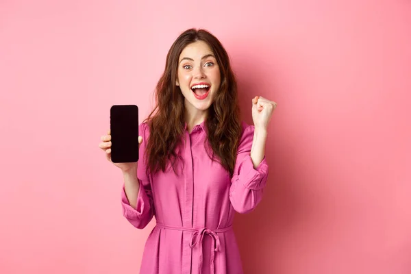 Technology concept. Excited girl shows mobile phone screen and scream with joy and happiness, winning money online, achieve daily goal on smartphone app, pink background