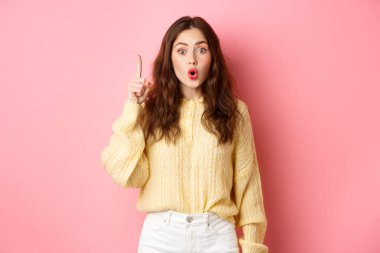 Heyecanlı genç esmer kız bir fikir sunuyor, parmağını kaldırıyor ve hayrete düşüyor, plan yapıyor, çözüm buluyor, pembe arka planda duruyor.