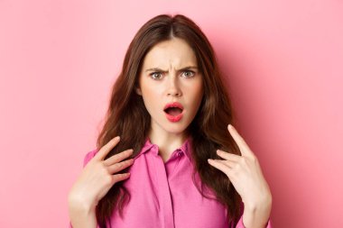 Kırgın ve şok olmuş genç bir kadına yakın çekim, pembe arka planda duran, kameraya somurtkan ve aşağılanmış bakışlar.