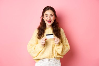 Plastik kredi kartıyla alışverişe çıkan heyecanlı kız kameraya şaşırmış bir şekilde gülümsüyor, pembe arka planda duruyor.