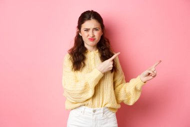 Şüpheci ve hoşnutsuz genç bir kadın somurtuyor, somurtuyor, somurtuyor ve parmaklarını telif alanına doğrultuyor, hoşnutsuzluğunu ve hayal kırıklığını dile getiriyor, pembe arka plana karşı duruyor.