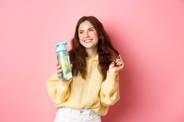 Güzel, çekici bir kadın saçlarıyla oynuyor ve gülümsüyor, yan bakıyor, su şişesinden sağlıklı bir içecek içiyor, pembe arka planda duruyor.