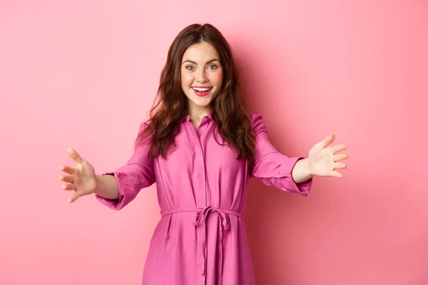 Stylish feminine lady reaching hands, stretched out arms to welcome friend, wants to hug or invite someone, standing against pink background