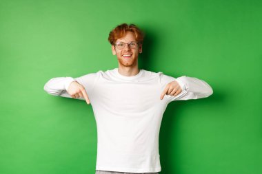 Kızıl saçlı, gözlüklü ve uzun kollu yakışıklı bir erkek model, parmaklarını aşağı uzatıp gülümsüyor, tanıtım yapıyor, yeşil arka planı var.